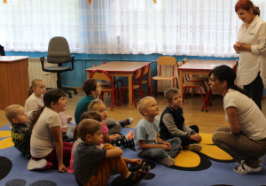 Przedszkolaki Miłosz, Lena, Maja, Olek, Denisa, Adaś, Hana i Karol siedzą na dywanie, przed nimi pani Kinga i pani Ula